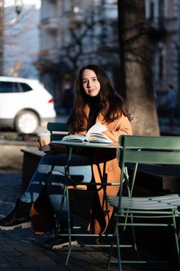 Genç bir kadın, arka planda şehir hayatı tarafından vurgulanan, sıcak güneş ışığı sahneyi aydınlatırken, kahvesini yudumlarken, kitaplarına dalmış bir şekilde bir kafenin dışında dinleniyor..