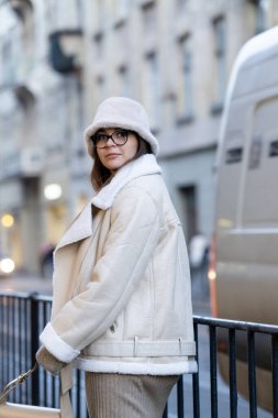 Şık, büyük bir ceket giymiş, gözlüklü bir kadın şehir kaldırımında yürüyor, şehir mimarisinin arka planına karşı şık bir hava yayıyor..