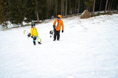 İki snowboardcu renkli kış kıyafetleri içinde karlı bir yamaca tırmanıyor, dingin bir ortamda dağdan aşağıya doğru heyecanlı bir yolculuğa hazırlanırken serin havanın tadını çıkarıyorlar..