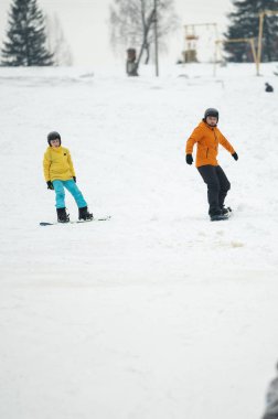 İki çocuk renkli snowboardlara taze kar oyuyor, neşe ve heyecan saçan kış harikalar diyarında karlı bir tepede gezinirken kahkahalar atıyorlar..