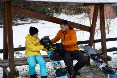Parlak sarı ceketli bir çocuk, snowboard ekipmanlarında rehberlik alır. İkisi de karlı bir ortamda ahşap bir bankta oturan turuncu ceketli bir adamdan..