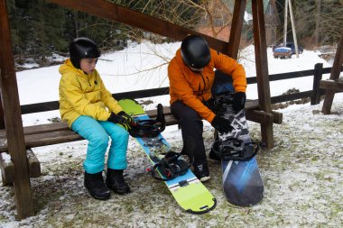 Genç bir hevesli ebeveyn, çam ağaçları ve kış sporları için mükemmel sıcak bir atmosfer ile çevrili karlı bir ortamda donanıma yardım ederken hevesle oturur..