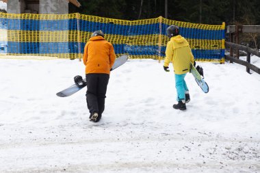 Canlı kış kıyafetleri giymiş iki snowboardcu kaykaylarını taşıyarak karlı arazide yürür renkli bir dağ tatil köyündeki yamaçlara hevesle yaklaşır..