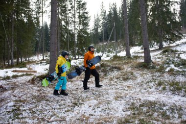 İki snowboardcu, parlak kış kıyafetleri içinde, karlı bir orman yolunda ilerliyorlar, heyecanlı bir snowboard günü için yamaçlara doğru ilerliyorlar..