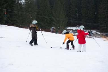 Parlak kayak kıyafeti giymiş üç çocuk kayak yeteneklerini karlı bir tepede geliştirmeye odaklanmış. Etrafı yemyeşil ağaçlarla çevrili. Kahkahalar kışın serin havasını dolduruyor..
