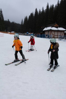 Üç çocuk kar yamaçlarında kayak yapıyor, kış için sıcak kıyafetler giyiyorlar. Arka planda uzun ağaçlar ve büyüleyici bir pansiyon yer alıyor ve güzel bir kayak merkezi atmosferi yaratıyor..