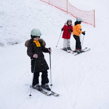 Çocuklar ilk kayak derslerini renkli kıyafetler ve miğferlerle karlı bir yamaçta süzülerek alırlar. Kahkaha ve eğlence kayak merkezindeki kışın serin havasını doldurur..