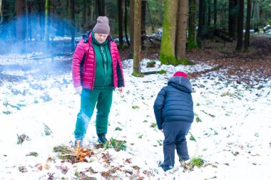 Bir yetişkin ve bir çocuk karlı bir ormanda küçük bir ateşe bakıyor. Sahne, kış macerası ve sessiz bir arkadaşlığı çağrıştırıyor..