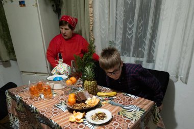 Kırmızı kazaklı yaşlı bir kadın ve gözlüklü genç bir çocuk meyve, şeker ve içeceklerle dolu bir masada oturuyorlar. Küçük bir Noel ağacı evlerindeki şenlikli atmosfere katkı sağlar..