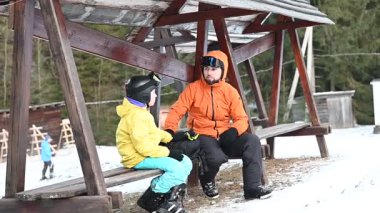 Sıcak kış giysileri içinde bir gün kayak yaptıktan sonra ahşap bir bankta dinlenen bir adam ve çocuğun yürek ısıtan bir sahnesi. Karlı kış manzarası onları kuşatıyor..