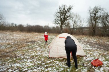 Soğuk bir çayırda, iki kişi çıplak ağaçların ve bulutlu bir gökyüzünün önünde kış keşfi ve aile bağlarının ruhunu kucaklayan bir çadır kurar..