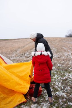 Kırmızı paltolu bir çocuk, serin bir kış günü, karlı bir arazide, sarı bir çadır kuran bir yetişkine yardım eder, neşe ve beraberlik anları yaratır..