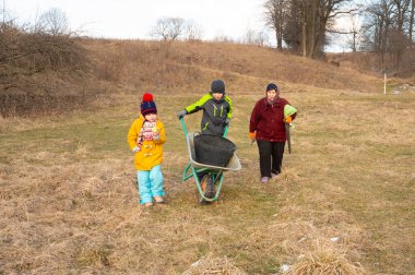 Çimenli bir alanda işbirliği yapan iki çocuk ve bir yetişkin, serin, bulutlu gökyüzünün altında içi ıvır zıvırla dolu bir el arabası taşırken takım çalışması sergiliyorlar..