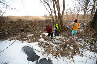 Üç çocuk, karlı bir arazide eğlenceli aktivitelere girişir. Ağaçlar arka planda dik dururken donmuş bir derenin kenarındaki çubukları toplar ve sakin bir kış ortamı yaratır..