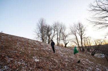 İki birey, güneşli kış alacakaranlıklarında çıplak ağaçlarla çevrili, yapraklarla ve hafif karla kaplı hafif bir yamaçtan geçer..