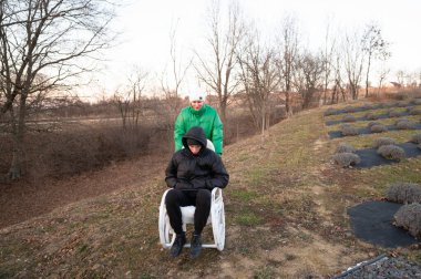 One friend pushes another down a gentle slope in a wheelbarrow, both dressed warmly for the cool weather, surrounded by bare trees and rows of lavender.