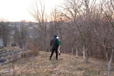 Güneş batarken çıplak ağaçlarla çevrili bir yamaç boyunca yürüyen iki birey serin akşam havasında sakin ve sakin bir atmosfer yaratıyor..