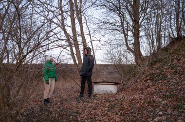 Alacakaranlıkta sakin bir ormanlık alanda çıplak dalların altında, iki arkadaş sohbet eder ve birbirleriyle eğlenirler, etrafları dökülen yapraklarla çevrilidir..