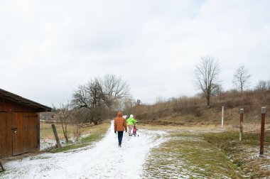 Aileler kış gezintisini severler, neşeli bir köpekle karlı bir patikada yürürler. Bulutlu gökyüzü bu serin araziye sakin bir atmosfer ekler.