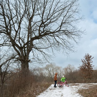 Parlak yeşil giyinmiş bir çocuk motosiklet üzerinde sevinçle mücadele ederken, bir yetişkin, karlı ve büyüleyici bir kış ortamında ayakta duruyor..