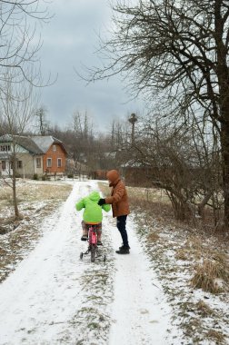 Serin bir kış gününde, parlak yeşil elbiseli bir çocuk rahat ceketli bir yetişkinin rehberliğinde ağaçlarla kaplı bir kır yolunda bisiklet sürmeye çalışıyor..