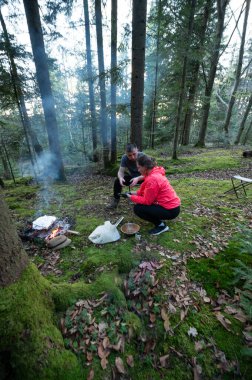 Bir çift, bereketli yeşil bir ormandaki küçük bir kamp ateşinin etrafında toplanır. Etrafı uzun ağaçlarla çevrilidir ve doğanın huzurunun tadını çıkarırlar..