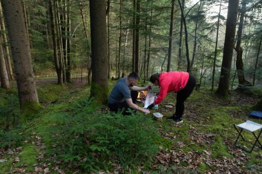 Sakin bir ormanda, iki kişi piknik hazırlıyor, battaniyeye yemek koyuyorlar. Güneş ışığı ağaçların arasından usulca parlıyor, huzurlu bir atmosfer yaratıyor..