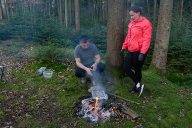 Huzurlu bir orman ortamında, bir çift, yüksek ağaçlar ve doğanın yatıştırıcı sesleri ile çevrili, açık bir ateş üzerinde lezzetli bir barbekü hazırlar..