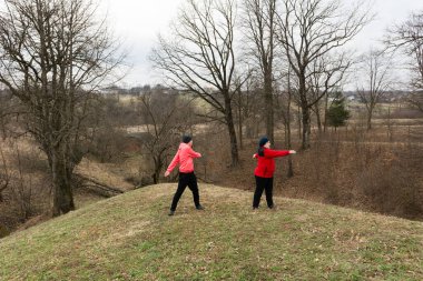 Alacakaranlık yaklaşırken iki insan çimenli bir tepenin üzerinde neşeyle dans ediyor. Yapraksız ağaçlar, sakin kırsal manzaranın geniş çerçevesini oluşturuyor..