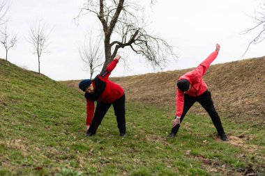 Kırmızı ceketli iki birey bulutlu bir gökyüzü altında çimenli bir yamaçta uzanıyor ve ilkbaharın başlarında doğanın huzuruyla çevrili sağlıklı bir sabah rutinini kucaklıyor..