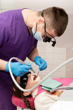 Yetenekli bir dişçi neşeli bir klinikte genç bir kızın dişlerini dikkatlice inceliyor. Kız güneş gözlüğü takıyor ve rahat görünüyor. Pratisyen ise özel aletler kullanıyor..