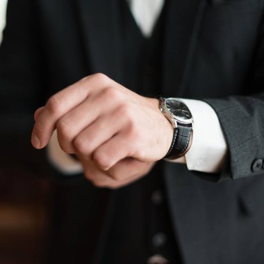 Kol saatini ayarlayan bir adamın elinin yakın çekimi. Saatte gümüş bir çanta ve siyah bir deri bant yer alıyor. Koyu renk bir takım giyiyor..