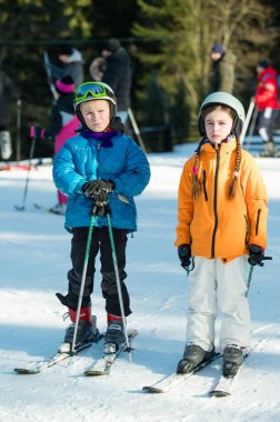 İki genç çocuk, kış meraklılarıyla dolu popüler bir kayak merkezinin karlı yamaçlarını keşfetmek için hevesle kayaklarının üzerinde duruyorlar..
