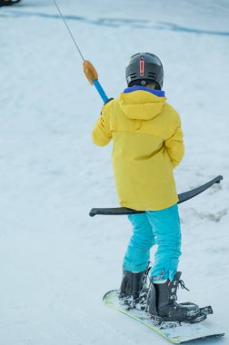 Canlı sarı bir ceket ve mavi pantolon giymiş genç bir kayakçı, güzel bir kış manzarasıyla çevrili bir kayak çekicisini tutarken karlı bir yamaçtan aşağı süzülür..