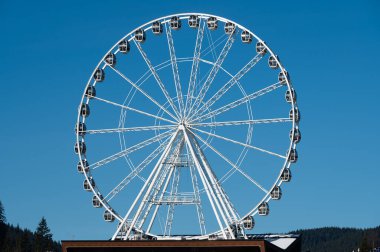 Parlak mavi bir gökyüzünün altında, yüksek bir gözlem çarkı sevinçle dönüyor, ziyaretçileri yakınlardaki yemyeşil ağaçların ve canlı park aktivitelerinin zevkli manzarasına davet ediyor..