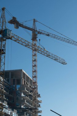 Ufuk çizgisine iki yüksek turna hükmediyor, çok katlı bir binanın inşaatını kolaylaştırıyor, işçiler aşağıdayken, şehir gelişimini iş başında gösteriyor.