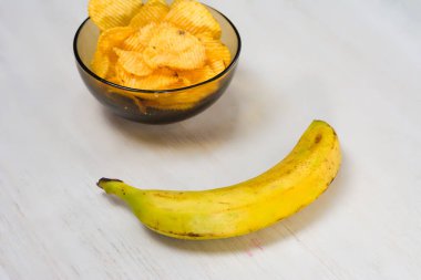 Beyaz ahşap bir yüzeye küçük bir cam kase dolusu patates cipsi ve olgun sarı bir muz yerleştiriliyor. Bu da basit bir atıştırmalık ya da hafif bir yemek olduğunu gösteriyor..