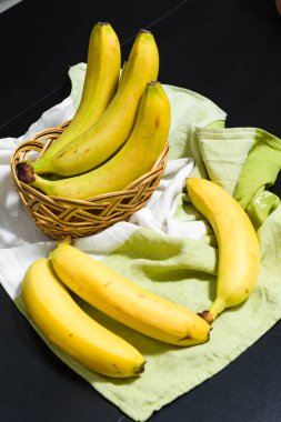 Bazı olgun sarı muzlar sergilenir, bazıları dokuma sepetinde ve diğerleri yeşil ve beyaz bir bez üzerinde dinlenir. Koyu arkaplan parlak sarı meyvelerle çelişiyor..