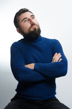 Mavi balıkçı yaka kazaklı sakallı bir adam kollarını kavuşturmuş, hafif bir arka plana bakarak yukarı bakıyor..