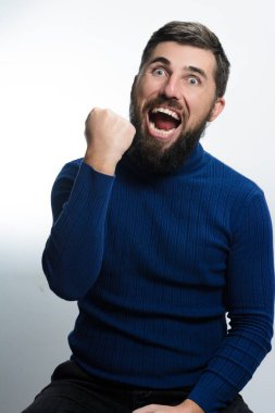 Mavi süveter ve siyah pantolon giyen sakallı bir adam beyaz bir arka plana yaslanır..