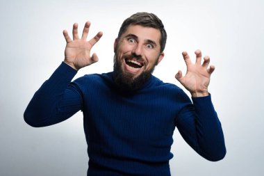 Mavi kazaklı sakallı bir adam kameraya doğru şakacı bir şekilde el hareketleriyle hareket eder. Yüzünde düz bir arkaplan karşısında komik bir ifade..