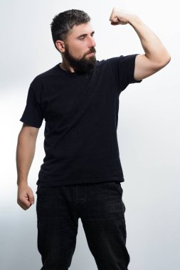 Siyah tişörtlü ve koyu renk kotlu sakallı bir adam kaslarını açık gri bir arka plana yaslar ve fiziksel gücünü ve kendine olan güvenini gösterir..