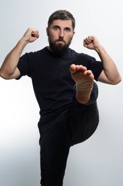 Siyah gömlek ve siyah pantolon giyen sakallı bir adam açık gri arka plana güçlü bir tekme atıyor. Yumrukları sıkılmış ve ifadesi odaklanmış..