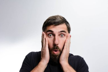 Sakallı ve koyu renk tişörtlü bir adam şok ve şaşkınlık gösterir. Elleri yüzünü ve gözlerini açık bir şekilde açık tutar..