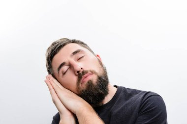 Kapalı gözlü sakallı bir adam, uykusunda ya da derin bir rahatlama jesti olarak, bembeyaz bir zemine yaslanır..