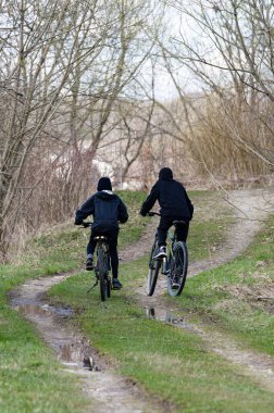 Bisikletli iki kişi toprak yolda gidiyor. Yol çamurlu ve tomurcuklanan ağaçlar ve yeşil çimlerle çevrili. Bisikletçiler rahat bir sürüşten hoşlanıyor gibi görünüyorlar..