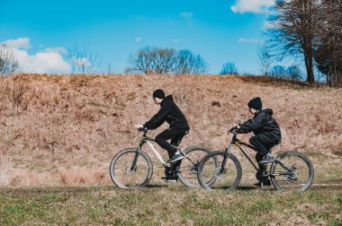 Dağ bisikleti süren iki kişi bir yol boyunca seyahat eder. Sahne, arka planında parlak mavi gökyüzünün altında bir tepe olan çimenli bir tarla..