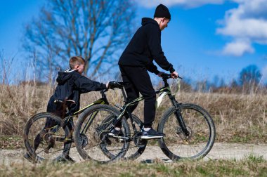 İki genç çocuk güneşli bir öğleden sonra bisiklet sürüşünün tadını çıkarıyor. Ağabeyi, küçük kardeşine toprak yolda yardım ediyor. Neşeli bir açık hava aktivitesi.