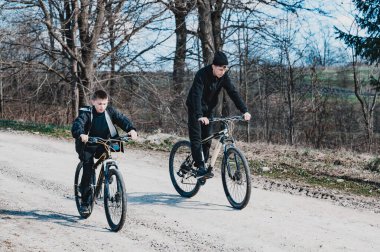 İki genç insan çakıllı bir patika boyunca bisiklet sürüyor. Ağaçlar yol boyunca uzanır, ve gökyüzü aydınlıktır, bir bahar ya da sonbahar gününü işaret eder. Sakin bir açık hava aktivitesi..