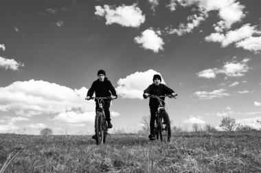 Bisiklet sürmekten hoşlanan iki gencin siyah beyaz fotoğrafı. Çimenli bir tarlada bisiklet sürerler, gökyüzü tüylü beyaz bulutlarla doludur. Özgürlük ve macera hissi uyandırıyor..